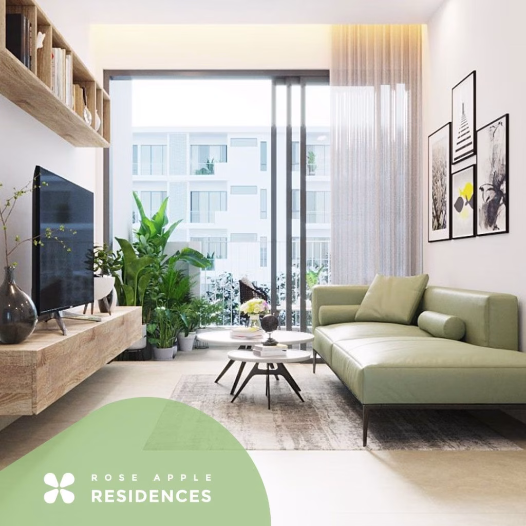 A modern living room at Rose Apple Square features a light green sofa, wooden coffee table, potted plants, wall art, a large TV on a wooden shelf, and floor-to-ceiling windows with views of an apartment building. Rose Apple Residences logo in the corner.