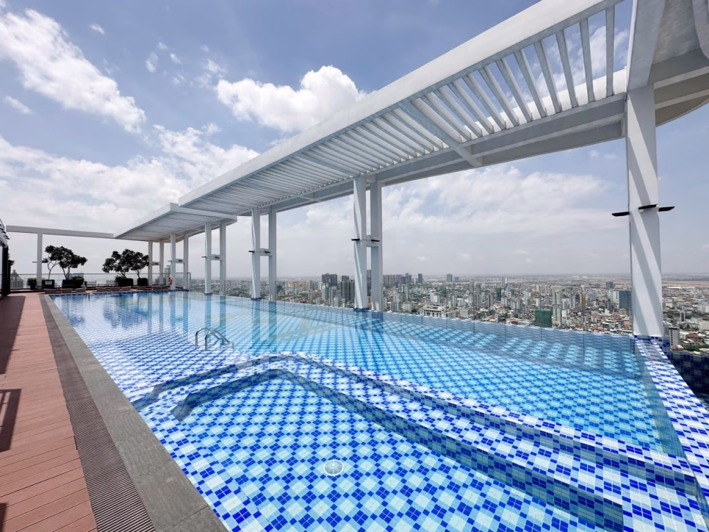 A rooftop infinity pool with blue and white tiles overlooks a city skyline under a partly cloudy sky. White pergola structures provide shade along one side, creating a relaxing retreat for Serviced Apartment 1 Bedroom residents.