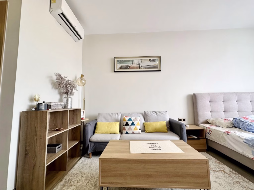 A cozy serviced apartment 1 bedroom living room with a gray sofa, colorful pillows, a wooden coffee table, and a bookshelf. A bed is partially visible on the right, and a landscape painting hangs above the sofa. An air conditioner is mounted on the wall.