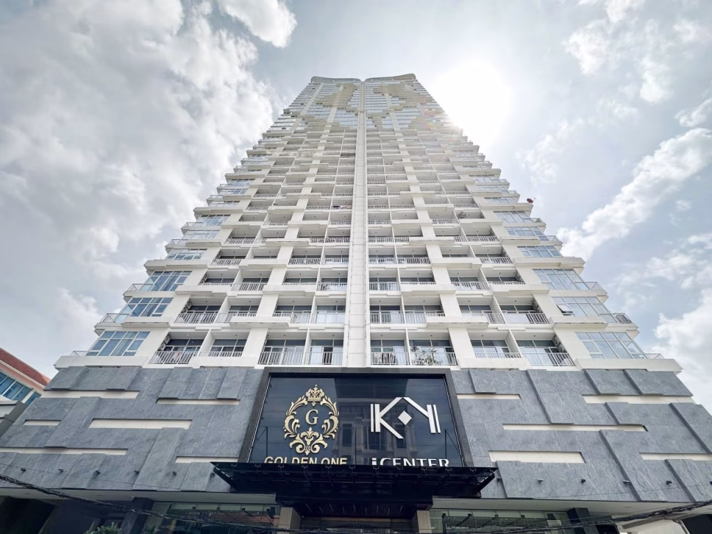 A tall, modern high-rise Serviced Apartment 1 Bedroom building seen from a low angle, with balconies on each floor and a decorative sign at the base reading GOLDEN ONE and KYN CENTER. The sun shines brightly in the partly cloudy sky above.