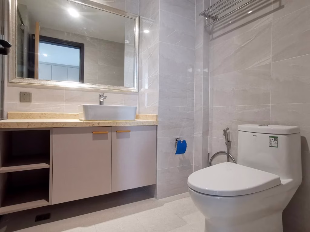 Modern bathroom at Star Bay Sihanoukville with beige tiles, a white sink on a light wood vanity beneath a large mirror, white toilet, and wall-mounted towel rack. The space appears clean and minimalistic.