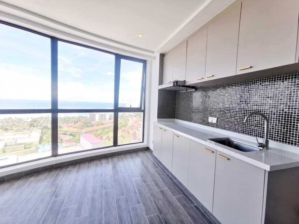 A modern kitchen with gray cabinets, a sleek countertop, black tile backsplash, and large floor-to-ceiling windows offering a panoramic view of Star Bay Sihanoukville’s cityscape and ocean.