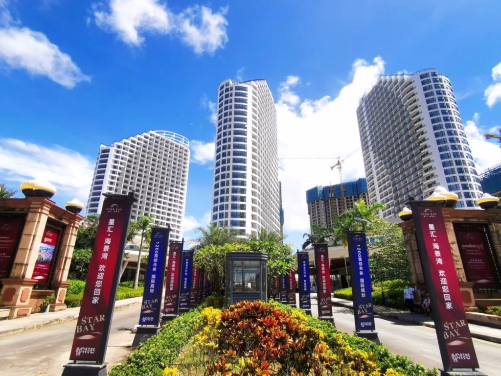 Tall modern buildings rise behind a landscaped walkway at Star Bay Sihanoukville, lined with colorful banners and lush plants under a bright blue sky with scattered clouds.