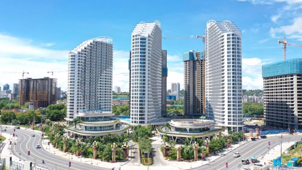 Aerial view of Star Bay Sihanoukville’s modern cityscape, featuring tall white and blue glass buildings, lush greenery, and curved roads under a bright sky. Construction cranes dot the skyline in the background.