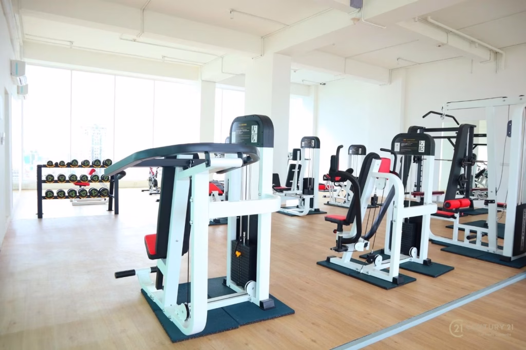 A bright, spacious gym—perfect for residents seeking fitness in comfort—features strength training machines, red and black padded seats, and a rack of dumbbells by sunlit windows near the studio apartment for rent.