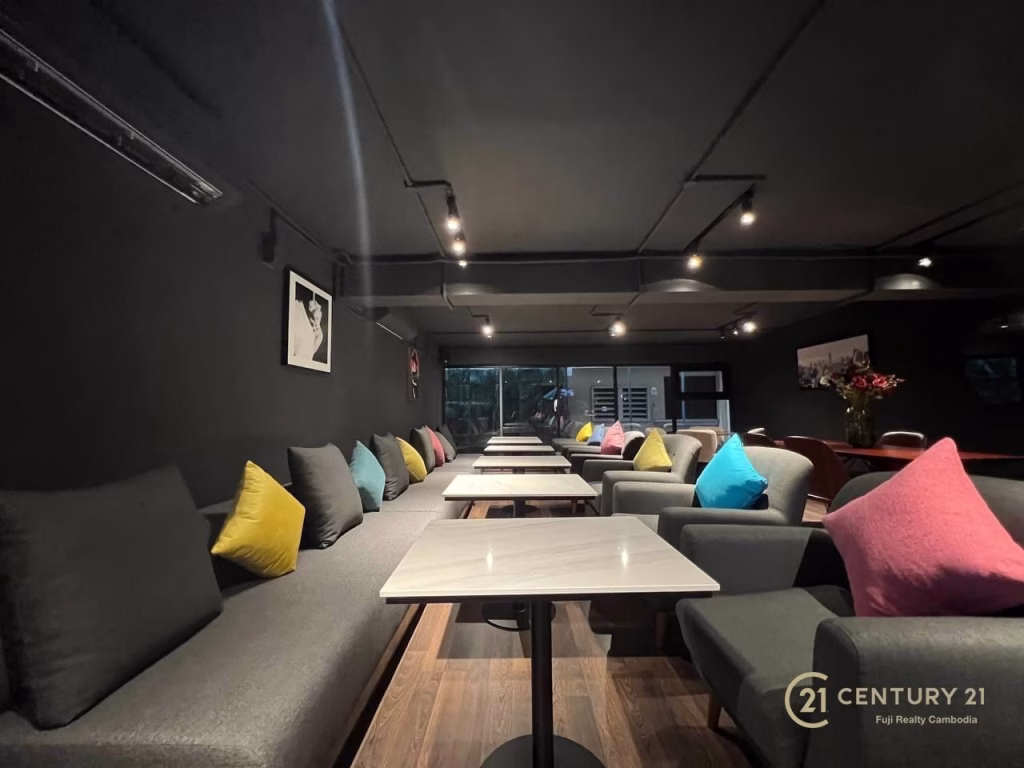 Modern lounge in a studio apartment for rent, featuring long gray sofas, colorful throw pillows, white tables, and dark walls. Framed art adorns the walls while large windows allow natural light. The Century 21 logo is visible in the bottom right corner.