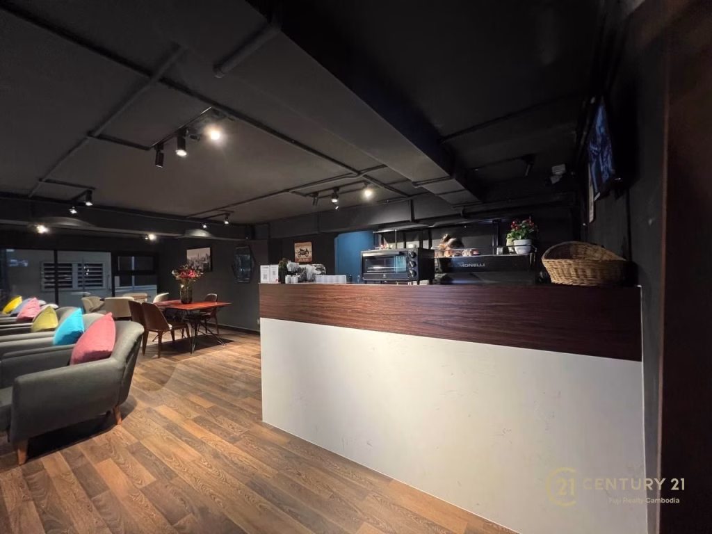 A modern café interior, perfect for a studio apartment for rent, with dark ceilings, wooden floors, a white and wood counter, sofas with colorful pillows, soft lighting, and flowers decorating the counter and tables.