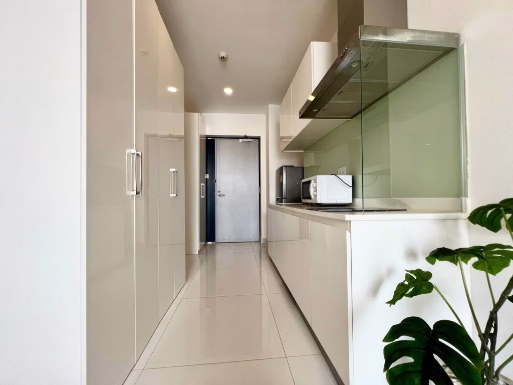 Modern, narrow kitchen in a studio apartment for rent features glossy white cabinets, built-in appliances, a microwave, and a glass backsplash; a green plant sits near the entrance door at the end of the hallway.