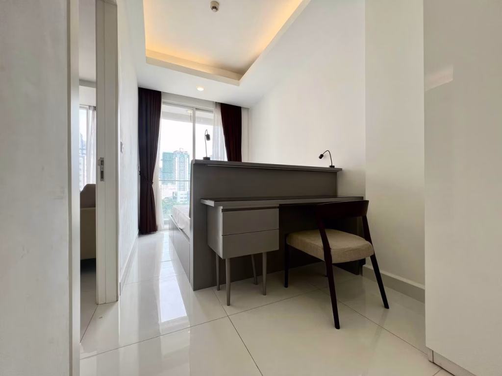 A modern, minimalistic studio apartment for rent featuring a small desk, chair, and lamp near a window with city view. White walls, glossy tiled floor, and dark curtains create a clean and bright atmosphere.