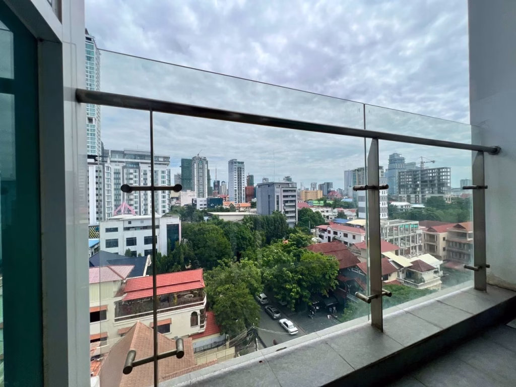 Cityscape view from a balcony with glass railing in a studio apartment for rent, showcasing high-rise buildings, mid-rise structures, and green trees under a cloudy sky. Several red-roofed houses are visible in the foreground.