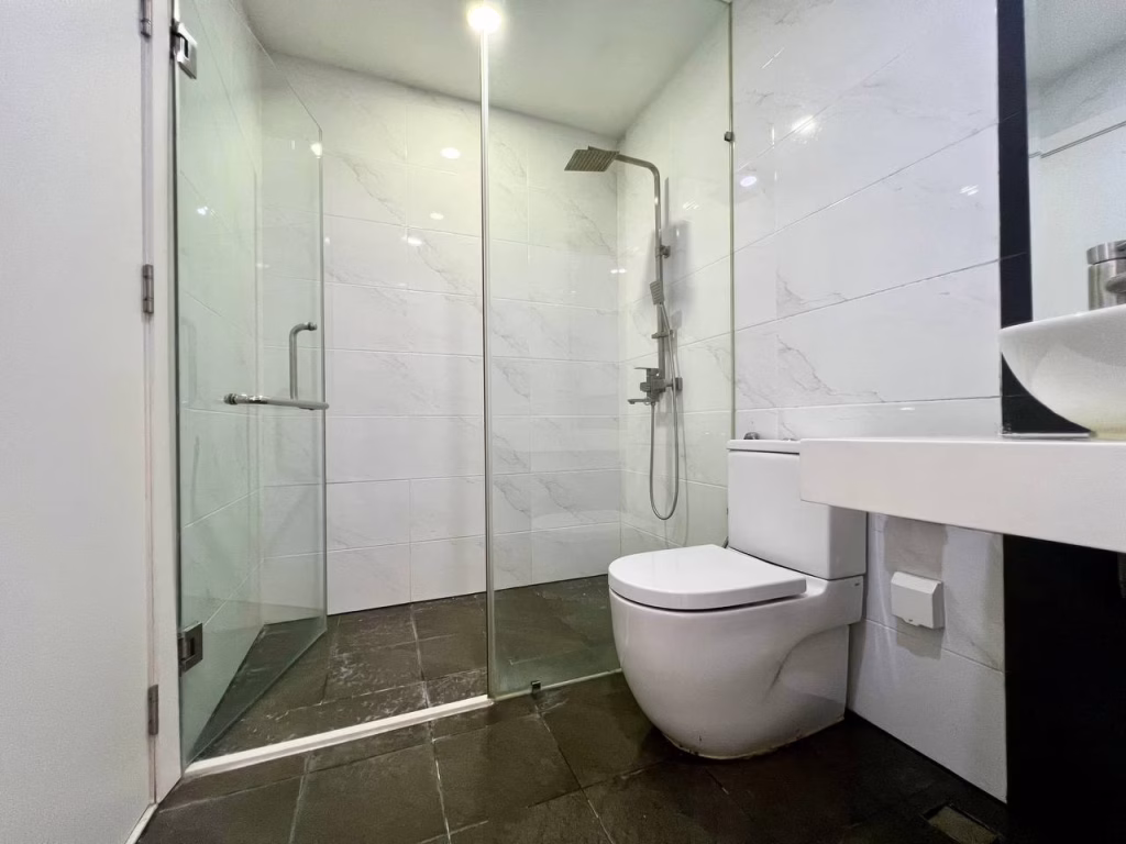 Modern bathroom in a studio apartment for rent with glass-enclosed shower, rainfall showerhead, white toilet, vessel sink on a countertop, white tiled walls, and dark floor tiles. The space is clean and minimalistic.