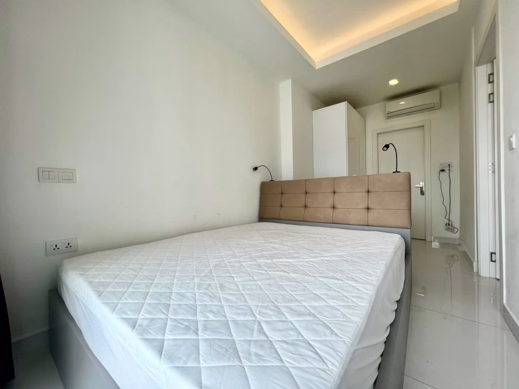 A modern, minimalistic bedroom in a studio apartment for rent features a neatly made double bed, padded beige headboard, white walls, built-in wardrobe, split air conditioner, and bright lighting from a recessed ceiling fixture.