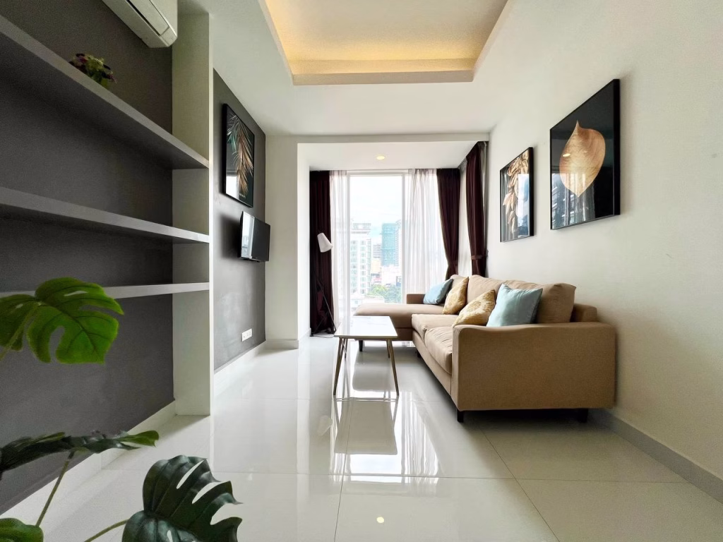 Modern living room in a studio apartment for rent, featuring a beige L-shaped sofa, decorative pillows, coffee table, and framed artwork. Large window with city view, potted plant, and wall-mounted TV create a bright, airy atmosphere.