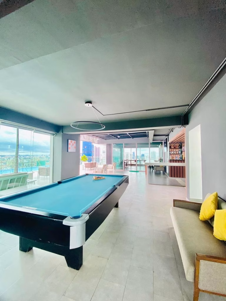 Stunning apartment for sale featuring a modern lounge with a pool table, gray sofa with yellow pillows, large windows flooding the space with natural light, and sleek décor—all set against a breathtaking city view.