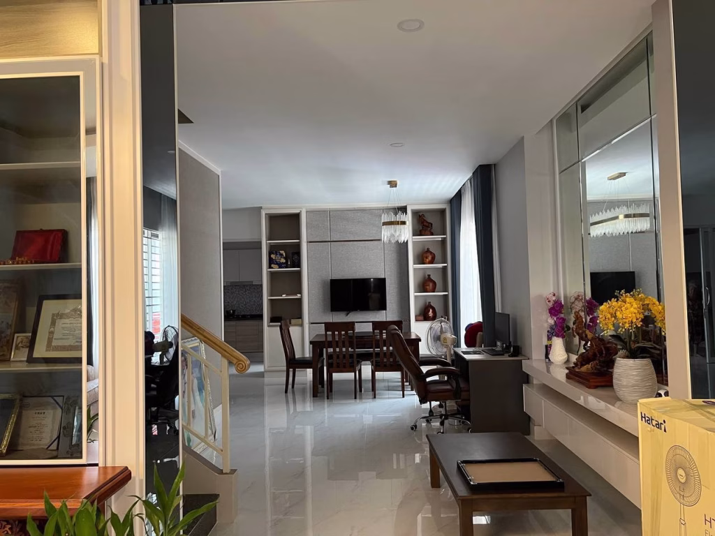 A modern living and dining area in this twin villa for sale features glossy white floors, a dining table, wall-mounted TV, decorative shelves, a desk with computer, plus various plants and decorative items for a stylish atmosphere.