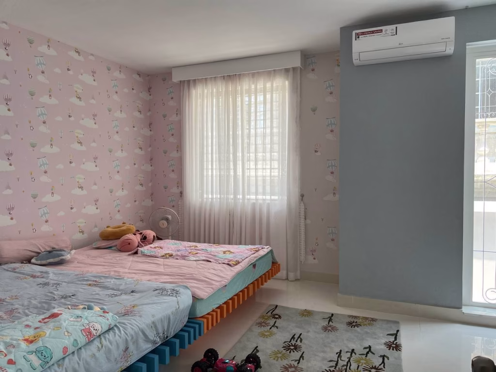 A bright bedroom in a twin villa for sale features two low beds with pastel bedding and pink whimsical wallpaper. Sunlight pours through sheer curtains, while an air conditioner cools the space and a rug with a toy car adds charm.