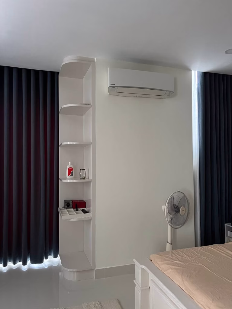 A minimalist bedroom in a twin villa for sale features dark curtains, a white wall-mounted air conditioner, a fan, a white shelving unit with a red mug and decor items, and a bed with a beige sheet.