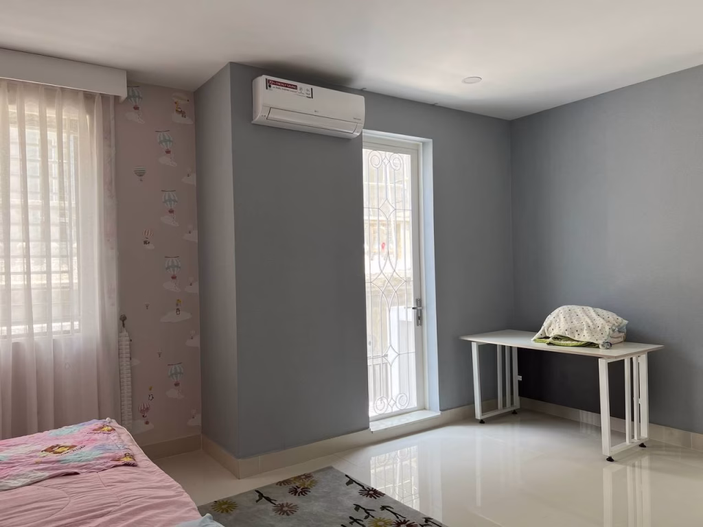 A bright, tidy bedroom in a twin villa for sale features pastel walls, an air conditioner, a window with sheer curtains, a single bed with pink bedding, a table with a pillow and bottle, and a door with a decorative metal grill.