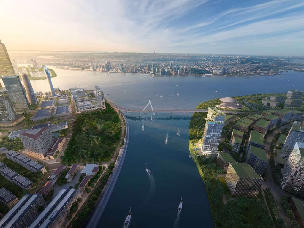 Aerial view of a modern cityscape with tall buildings, green parks, and riverfront luxury; a river runs through the center, boats glide along the water, and the opposite shore is visible under a blue sky.