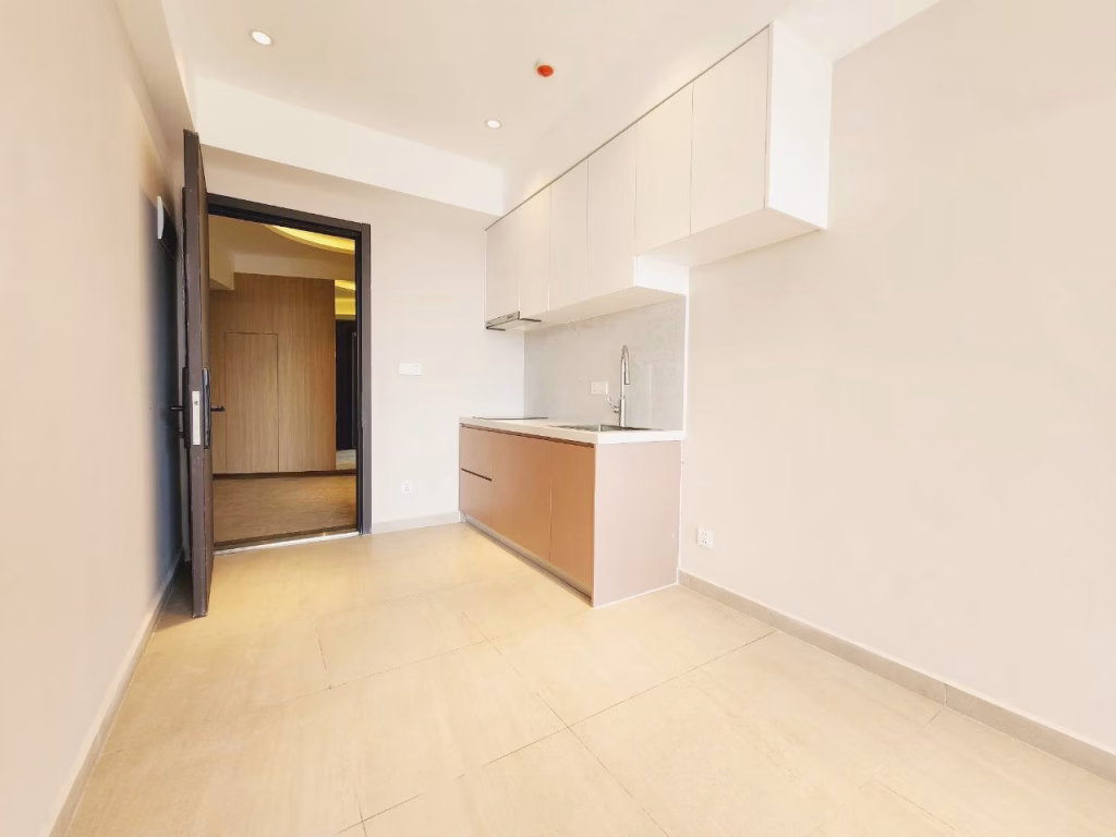 Minimalist, modern kitchen in a 1 bedroom condo with light beige floor tiles, white upper cabinets, light wood lower cabinets, and a marble backsplash. An open door leads to a warmly lit hallway. Room is empty and brightly lit.