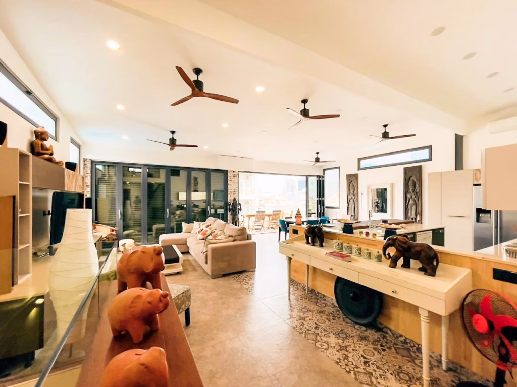 A spacious, modern living room in a 3-bedroom apartment features ceiling fans, large windows, neutral-toned furniture, animal figurines, and a patterned floor section. The area is well-lit with a mix of natural and artificial light.