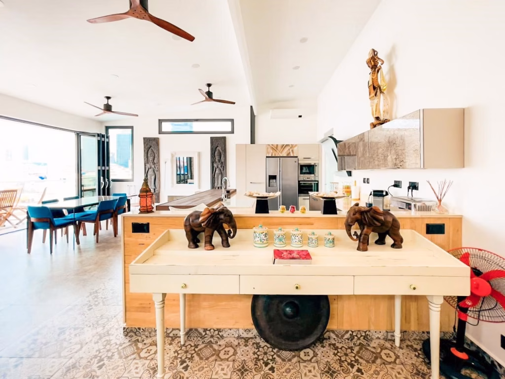 Modern, open-concept kitchen and dining area in a 3-bedroom apartment featuring wooden elephant sculptures, a gong, decorative jars, ceiling fans, and statues. Large windows fill the space with natural light; patterned tile floors and wood accents shine.