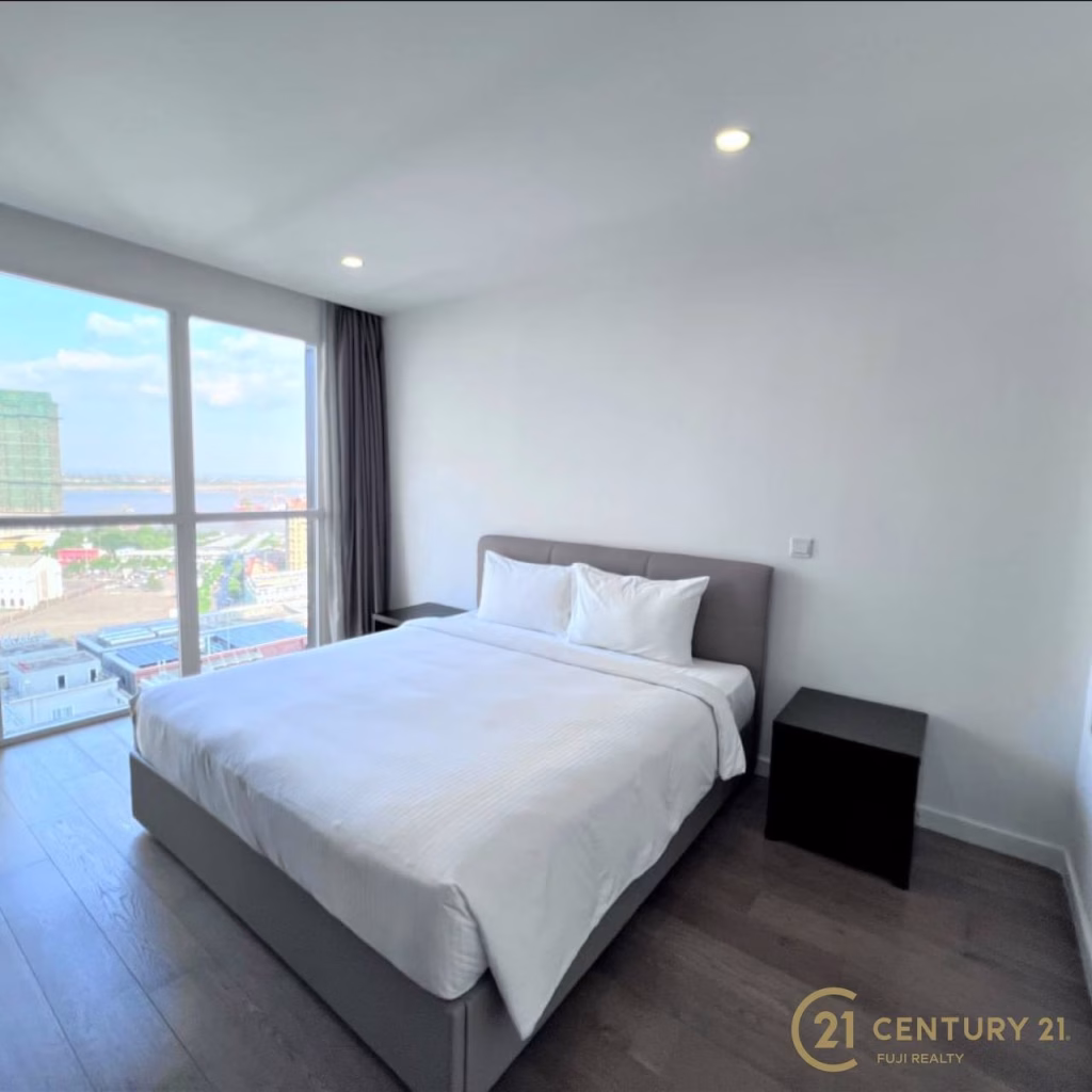 Modern bedroom at Meridian Koh Pich with a large bed, white bedding, two black nightstands, floor-to-ceiling window offering city views, and wood flooring. Logo for Century 21 Fuji Realty in lower right corner.