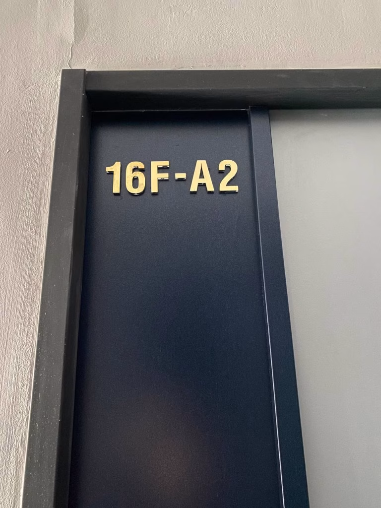Gold letters and numbers 16F-A2 are mounted on a dark blue door with a black frame in a condominium at BKK1. A light-colored wall is partially visible on the left side.