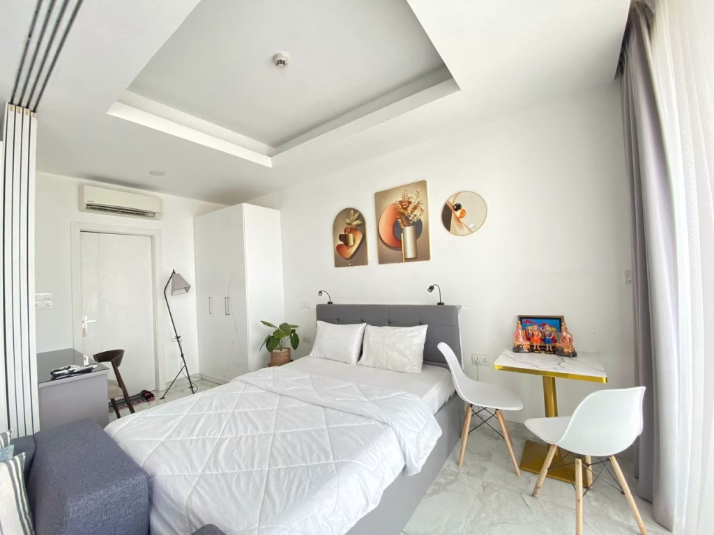 A bright, modern bedroom in a condominium at BKK1 features a neatly made bed, white walls, decorative art above the headboard, a small table with two white chairs, and large windows with sheer curtains letting in natural light.
