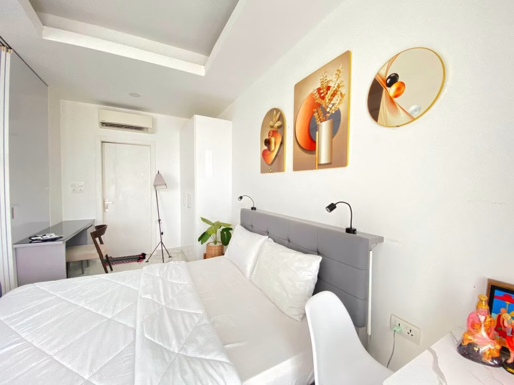 A bright, modern bedroom in a condominium at BKK1 features a neatly made bed with white bedding, a gray headboard, abstract art above the bed, plus a desk with chair, mirror, plants, and a small figurine in the corner.