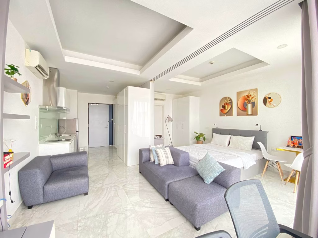 A modern condominium at BKK1 studio with gray sofas, a bed, small dining table, and minimalist decor. The room features white walls, marble floors, large windows for natural light, and a visible kitchen area in the background.