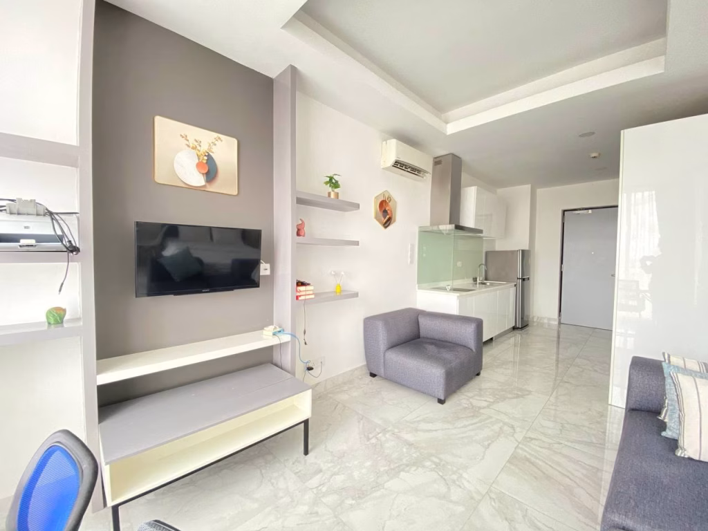 Modern, bright living space in a condominium at BKK1 with white and gray decor, a wall-mounted TV, shelves with small decor items, gray armchair, kitchenette, and marble flooring. Light fills the room from an unseen window.