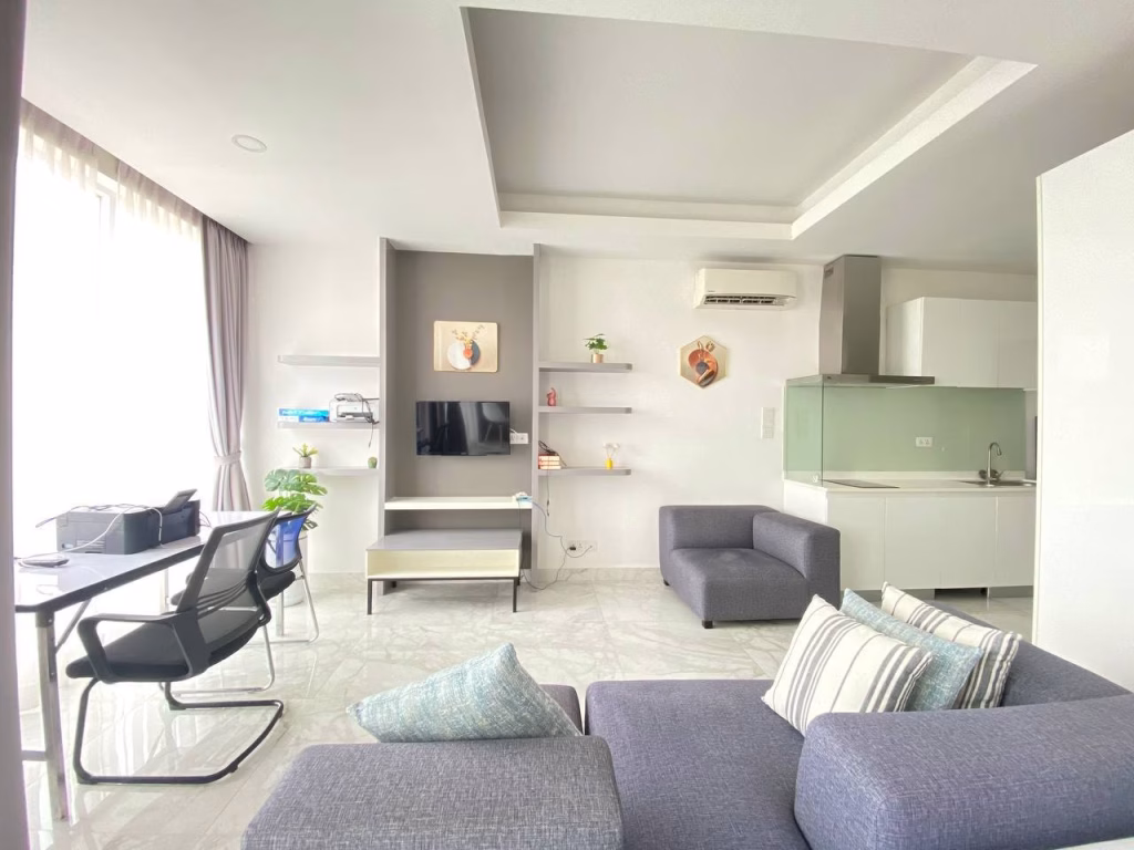 Modern living room in a condominium at BKK1 with gray sofa, pillows, and small table facing a wall-mounted TV. Desk with chair and plant by large window. Open kitchen features white cabinets, light green backsplash, and bright natural lighting.