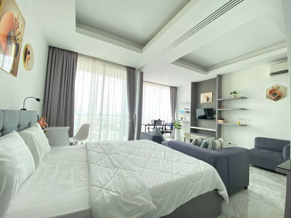 A modern, bright condominium at BKK1 features a neatly made bed, gray sofa, TV, shelves, desk area, and large windows with sheer curtains letting in natural light. Minimalist decor and soft tones create a cozy atmosphere.