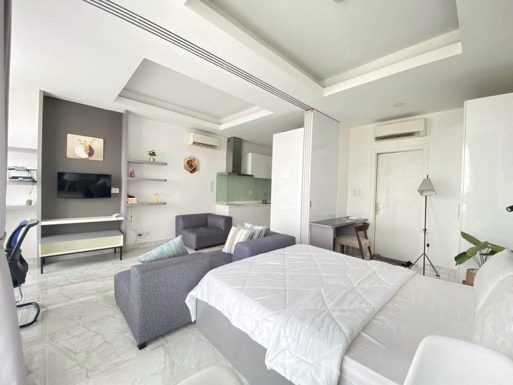 A bright, modern studio apartment in a condominium at BKK1 with a bed, gray sofa, desk, TV, shelves, and a kitchen in the background. The room features white walls, marble floors, and minimalistic decor.