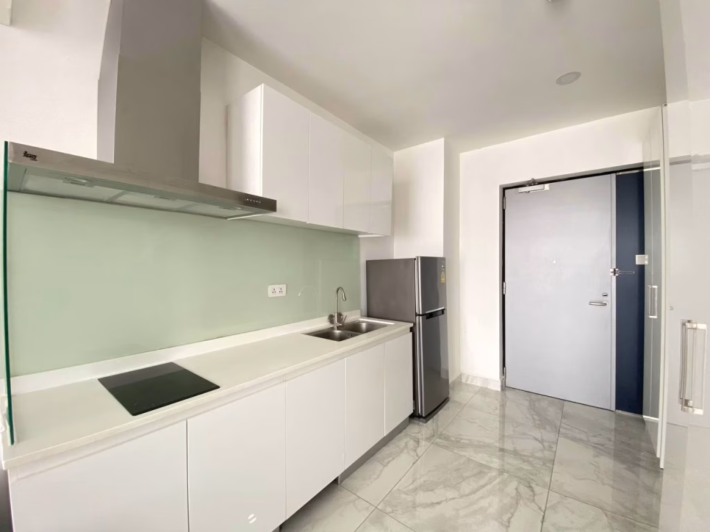 A modern kitchen in a condominium at BKK1 features white cabinets, an electric stove, a stainless steel sink, refrigerator, and light gray marble flooring. The walls are light, complemented by a sleek glass backsplash behind the stove.
