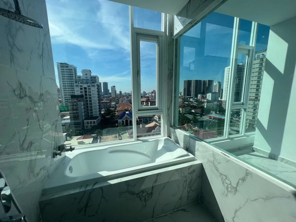 A modern bathroom with a marble bathtub by large windows in J Tower 2 Phnom Penh, offering a panoramic city view with tall buildings under a blue sky dotted with wispy clouds.