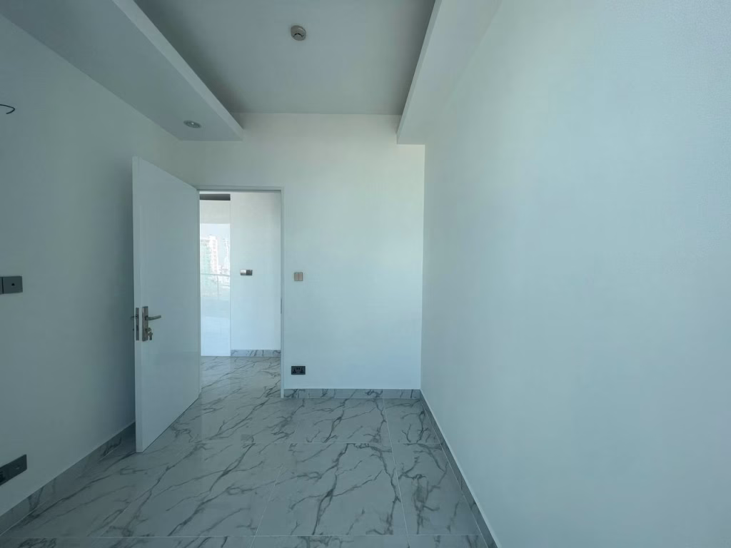 A small, empty room at J Tower 2 Phnom Penh features white walls and a door slightly open to a bright hallway. The floor is covered with grayish marble tiles, while sunlight streams in from another space outside the room.