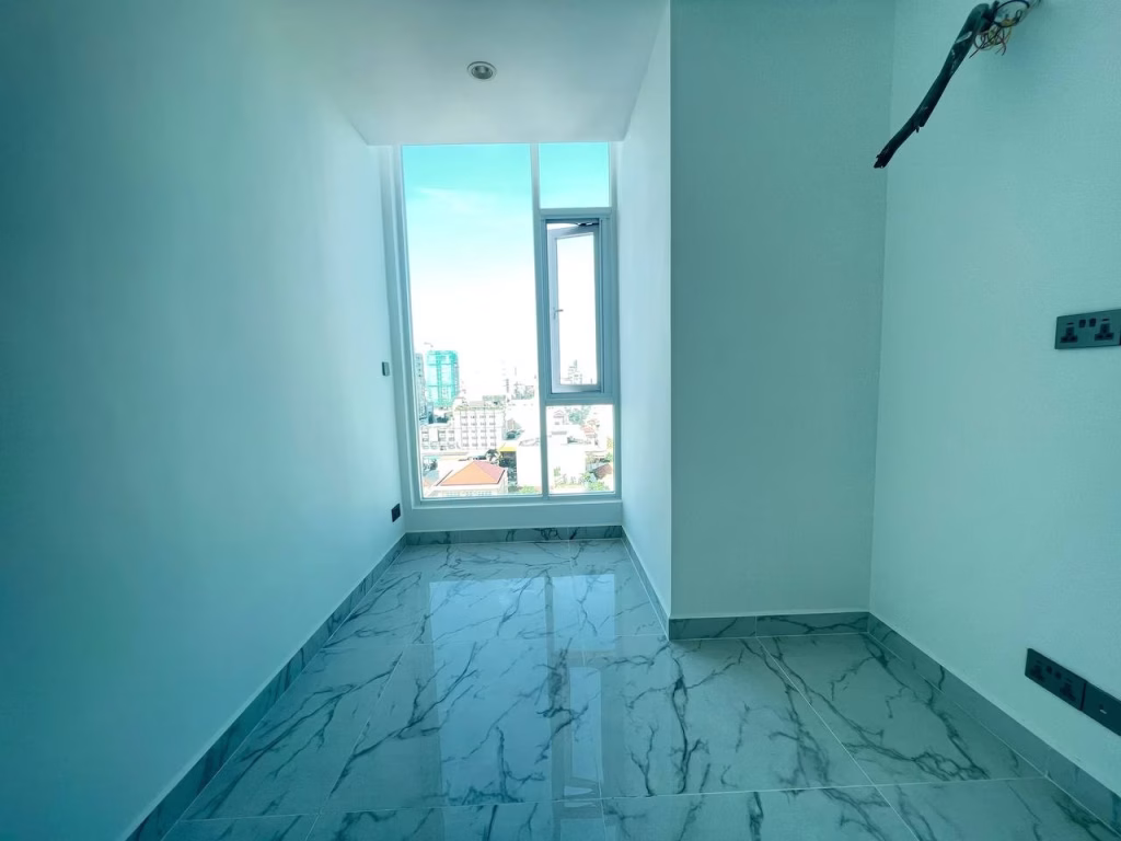 A small, empty room in J Tower 2 Phnom Penh features white walls and a marble tiled floor. A tall window lets in bright natural light and reveals the cityscape, while electrical outlets and a ceiling light fixture are visible.