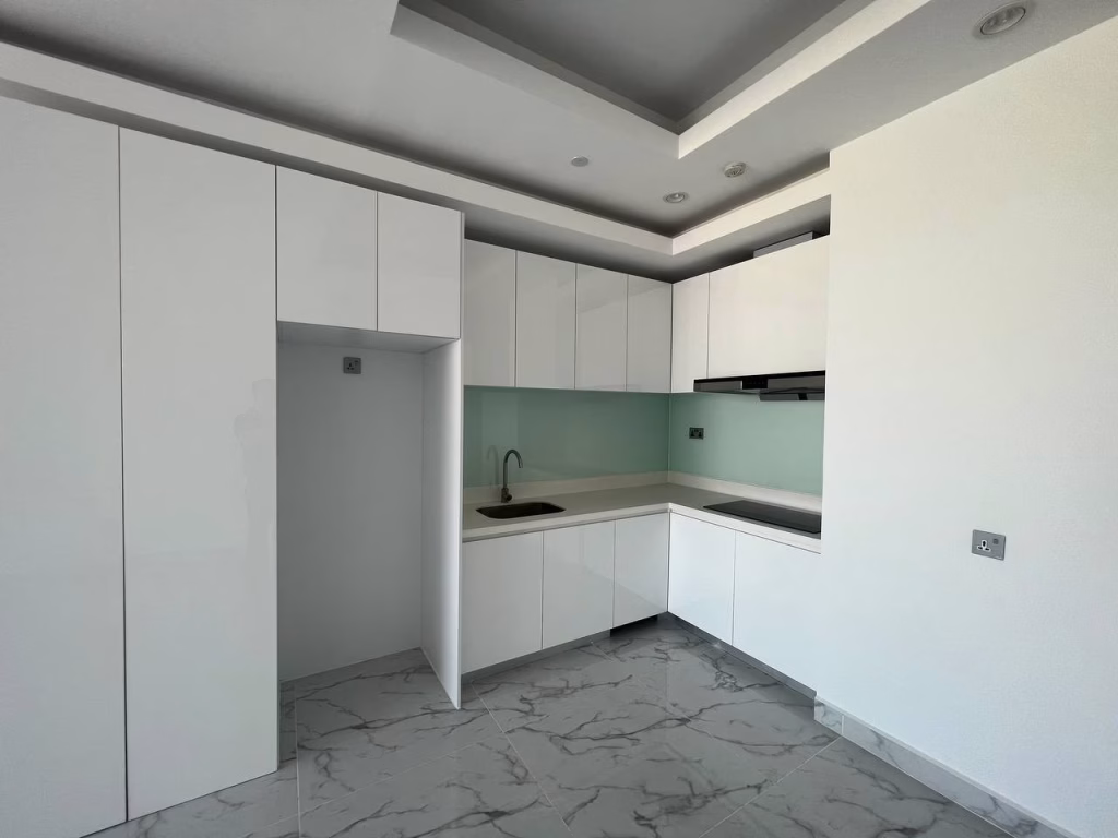 Minimalist, modern kitchen at J Tower 2 Phnom Penh with glossy white cabinets, sleek countertops, a built-in sink, stovetop, and a light green glass backsplash. White marble-tiled floors with gray veining complete this bright, uncluttered space.