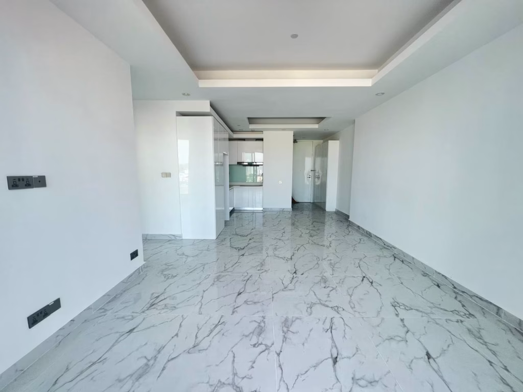 A modern, empty room in J Tower 2 Phnom Penh features white walls, a glossy gray marble floor, an open doorway to the kitchen area, and a ceiling with recessed lighting and a tray design.