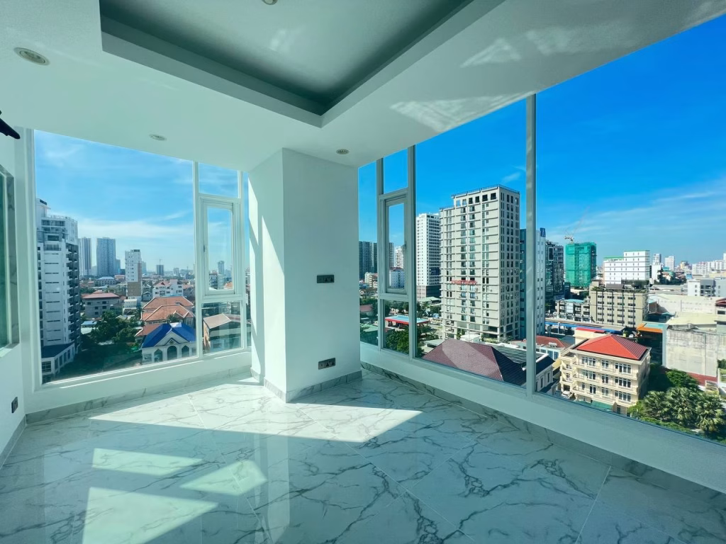 Bright, empty room in J Tower 2 Phnom Penh with large floor-to-ceiling windows offering a panoramic city view. Sunlight casts shadows on the white marble floor, and tall buildings are visible outside under a clear blue sky.