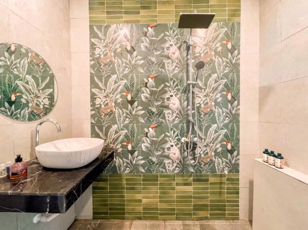 A bathroom in a villa for sale in Kampot features a black countertop and white vessel sink, a round mirror above, and a shower area with green tiles and tropical leaf-patterned wallpaper adorned with animals and birds.