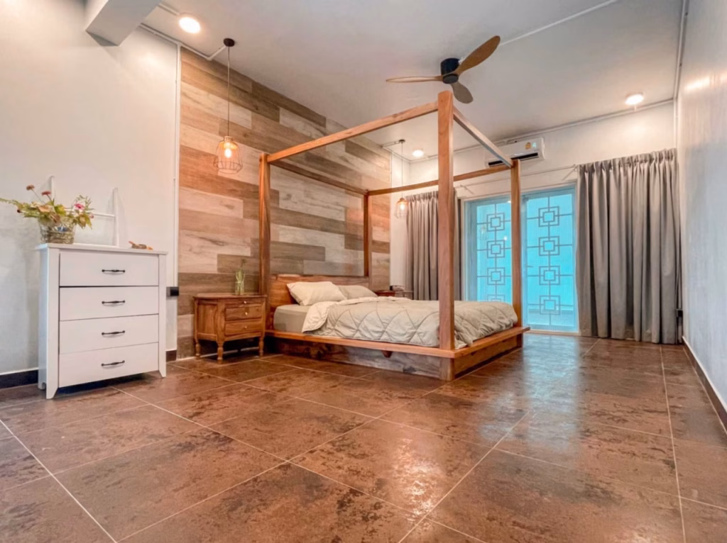 A spacious bedroom in a villa for sale in Kampot features a wooden canopy bed, neutral bedding, wood-paneled accent wall, ceiling fan, bedside table, white dresser with a flower vase, large tiled floor, and curtains over a windowed door.