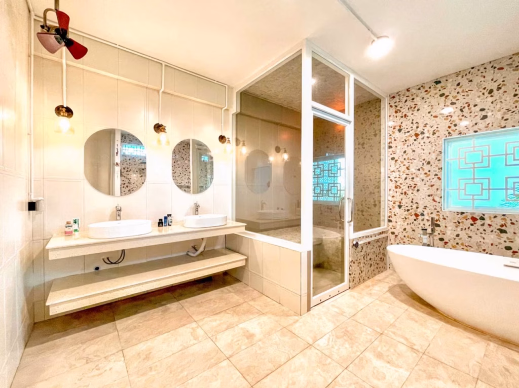 Modern bathroom in a villa for sale in Kampot, featuring two round mirrors and sinks on a floating counter, open shelves below, a large glass shower, freestanding bathtub, terrazzo accent wall, tiled floor, and warm lighting.
