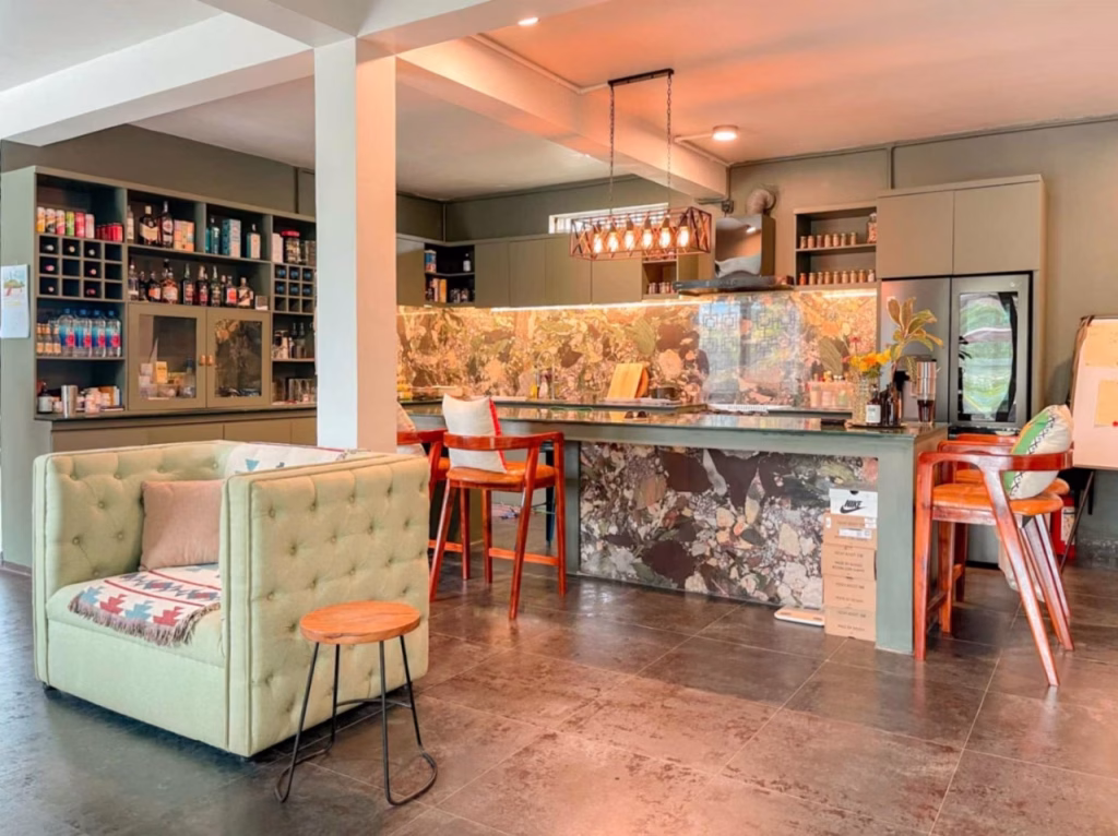 A modern, cozy café interior with a marble-patterned bar, wooden stools, a sage green sofa, and shelves stocked with various products and bottles—perfect inspiration for your dream villa for sale in Kampot. Warm lighting creates an inviting atmosphere.