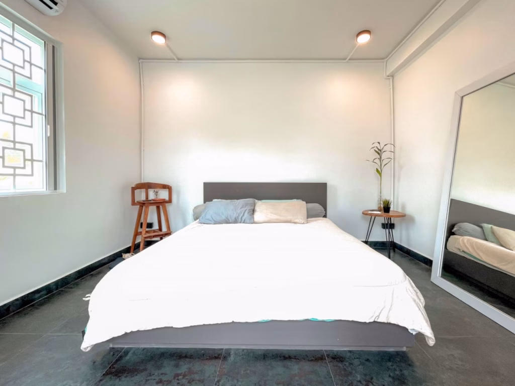 Minimalist bedroom in a villa for sale in Kampot, featuring a large bed with white bedding, two pillows, a wooden side table with a plant, a mirror on the right, and barred window on the left. Light walls contrast with dark flooring.