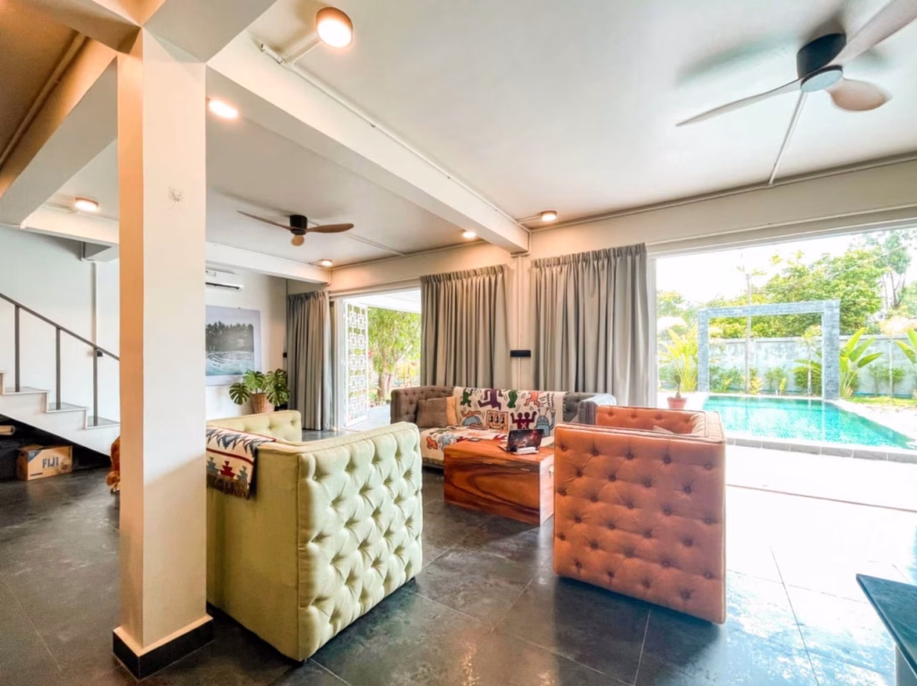 Modern living room in a villa for sale in Kampot, featuring stylish armchairs, a sofa facing a wooden coffee table, ceiling fans, large windows with curtains, and stunning views of the swimming pool and lush greenery outside.