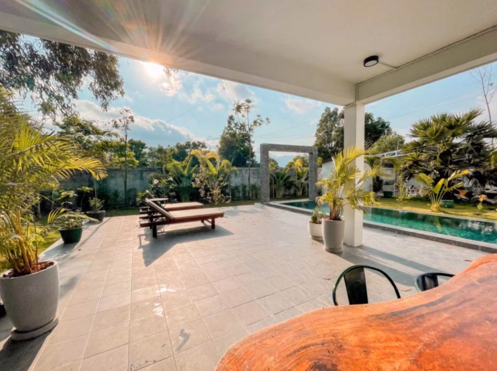 A sunny patio with potted plants, lounge chairs, and a swimming pool surrounded by greenery. Sunlight streams in, casting shadows across the tiled floor of this villa for sale in Kampot under a covered area with a wooden table.