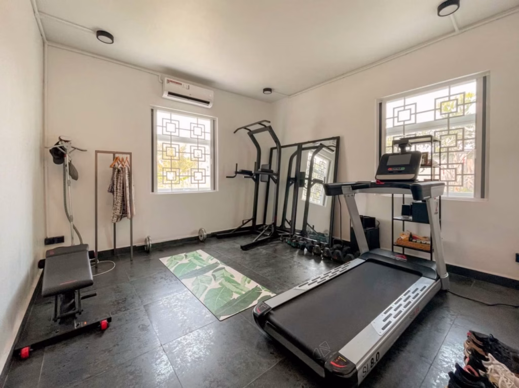 A villa for sale in Kampot features a home gym with a treadmill, weight bench, exercise mat, dumbbells, mirrors, and a clothes rack set against white walls and two windows with barred grilles. The floor is tiled in dark gray.